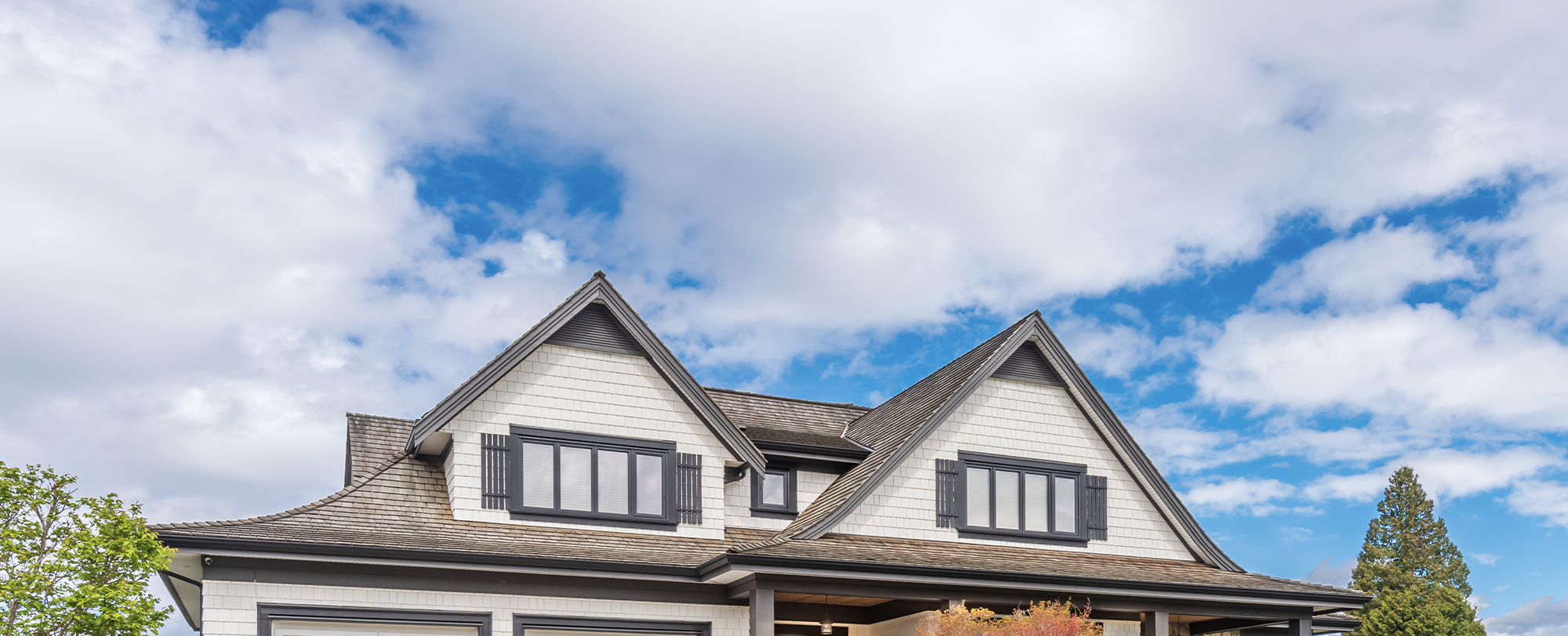 Tan shingled house with dark trim and shutters against a blue sky